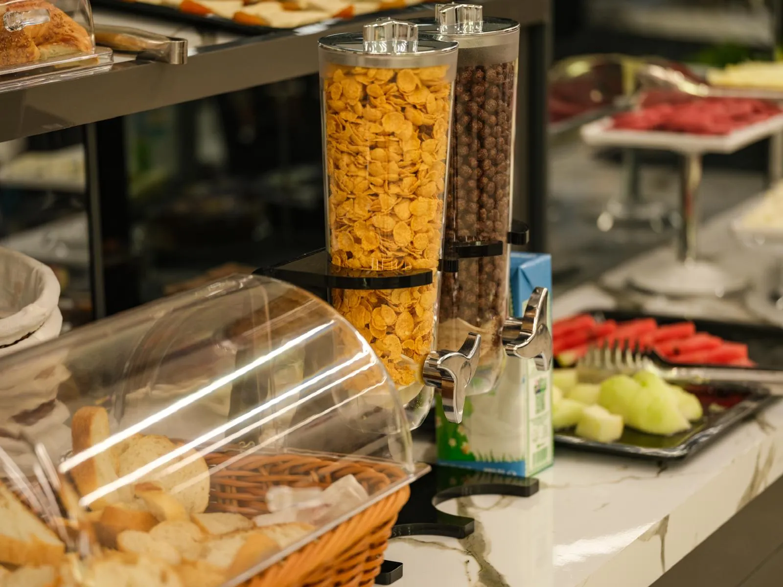Kahvaltı büfesinde mısır gevreği dispenserleri, ekmek sepeti ve dilimlenmiş karpuz